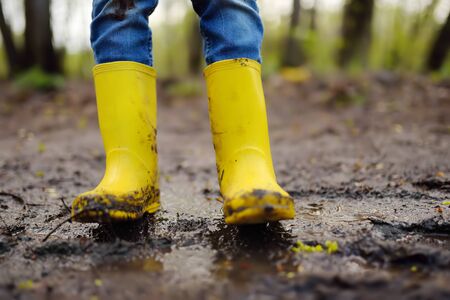 Mischievous preschooler child wearing yellow rubber rain boots jumping in large wet mud puddle. Kid playing and having fun. Outdoors games for children.の写真素材