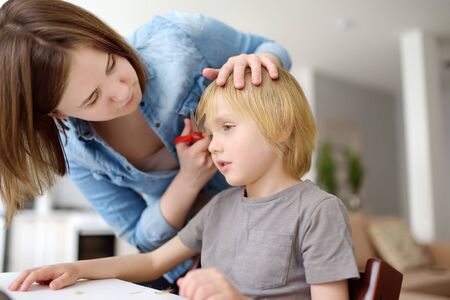 Preschooler boy gets haircut at home during quarantine. Mother cuts his son's bangs with scissors and comb. Hair cutting for kids by parents while hairdressers closed.の写真素材