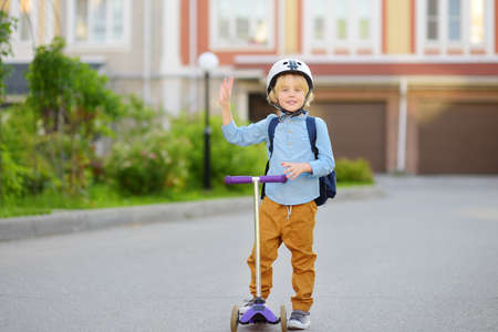 Little child in safety helmet riding scooter to school. Preschooler boy waving hand saying hi. Safety kids by way to school.の写真素材