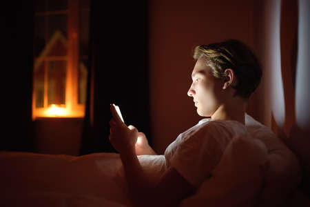 Teenager boy chatting by phone, writing in blog or reading social networks instead sleeping. Young...