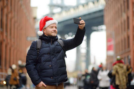Middle aged man tourist in Santa Claus hat takes selfie on street near the Manhattan Bridge in New...