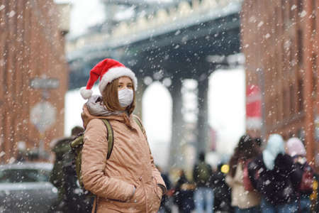 Young local woman in Santa Claus hat walks during a snowfall in New York City on Christmas Eve. Face mask and social distancing - safety in public place on winter holidays while coronavirus epidemic.の写真素材