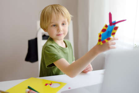 Little boy making colored paper crafts at home during coronavirus lockdown. Child showing result...