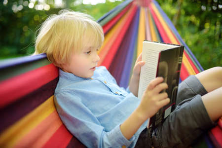 Cute little blond caucasian boy reading book and having fun with multicolored hammock in backyard or outdoor playground. Summer active leisure for kids. Child swinging and relaxing in hammock.の写真素材