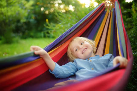 Cute little blond caucasian boy enjoy and having fun with multicolored hammock in backyard or outdoor playground. Summer outdoors active leisure for kids. Child relaxing and swinging in hammock.の写真素材