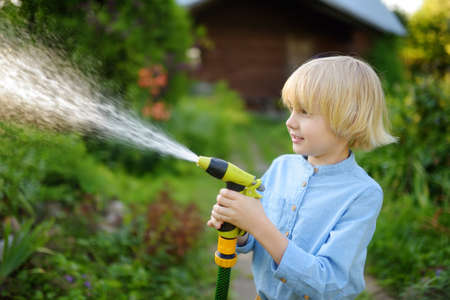 Funny little boy watering plants and playing with garden hose with sprinkler in sunny backyard. Preschooler child having fun with spray of water. Summer outdoors activity for kids.の写真素材