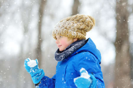 Little boy having fun playing with fresh snow during snowfall. Snowball fight. Kid dressed in warm...