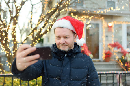 Mature man wearing Santa hat making selfie near his decorated home. Happy owner decorated his house and yard with garlands for Christmas and New Year.の写真素材