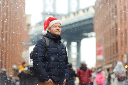 Middle aged man tourist in a Santa Claus hat is walking during a snowfall near Manhattan Bridge in New York on a snowy Christmas Eve. Winter Xmas holidays in NY. New Year vacations in NYC.の写真素材