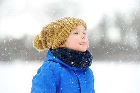 Cute child wearing a warm hat catching snowflakes with his tongue. Funny little boy in blue winter clothes walks during a snowfall. Outdoors winter activities for kids.の写真素材