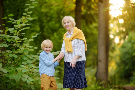 Cute grandson holding hands with his joyful elderly grandmother during walking at summer park. Two generations of family spend time together. Quality family timeの写真素材