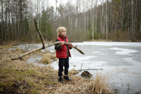 Little boy in red vest playing large branch on shore of forest lake on early spring day. Snow has just cleared, but surface of lake is still under ice. Outdoor activity for kids. Beauty of nature.の写真素材