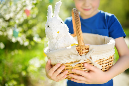Cute little boy holding wicker basket with white toy rabbit during hunt for colorful eggs in spring park on Easter day. Traditional entertainment for kids during easter festival outdoorsの写真素材