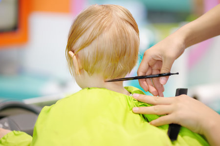 Preschooler boy getting haircut. Rear view. Children hairdresser with professional tools - comb and scissors. Cutting hair for kids.の写真素材