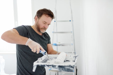 Portrait of a handsome mature man making repairs in the apartment. The person paints the wall white with roller. Do it yourself. DIY. Repair with their hands.の写真素材