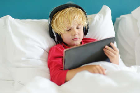 Cute preschool boy wearing headphones watches cartoon movie by tablet in bed in the morning right after waking up. Overuse and addiction kids from gadgets.の写真素材