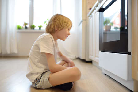 Glad boy sitting on the floor near kitchen stove and waiting for a pie or other baked goods to be prepared. Children love food prepared by their mother. Domestic cuisine. Healthy food for kids.の写真素材