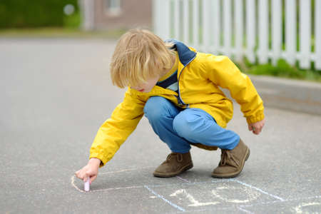 Little kid boy drawing hopscotch with colored chalk on asphalt. Child playing hopscotch game on playground outdoors on a spring day. Outdoor activities for children.の写真素材
