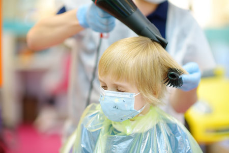 Preschooler boy wearing facemask getting haircut in salon during coronavirus epidemic. Children hairdresser drying hair of child with hair dryer. Cutting hair for kidsの写真素材