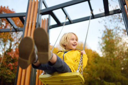 Little boy having fun on a swing on the playground in public park on autumn day. Happy child enjoy...