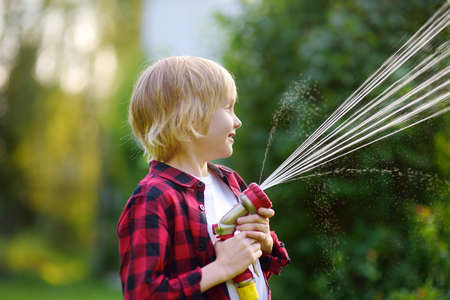 Funny little boy watering plants and playing with garden hose with sprinkler in sunny backyard. Preschooler child having fun with spray of water. Summer outdoors activity for kids.の写真素材