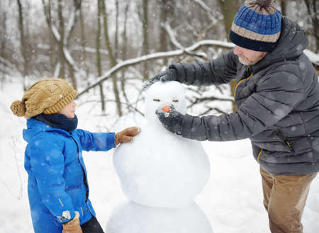 Little boy with his father building snowman in snowy park. Active outdoors leisure with family with children in winter. Kid during stroll in a snowy winter parkの写真素材