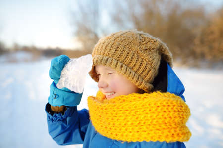 Little boy having fun playing with icicle in forest on sunny winter day. Baby looking through ice at sun. Active leisure outdoors for child on nature among driven snowの写真素材