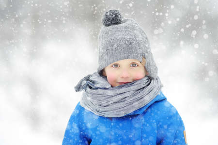 Portrait of funny little boy in blue winter clothes walks during a snowfall. Cute child wearing a warm clothing, hat, scarf is looking at you. Outdoors winter activities for family with kids.の写真素材