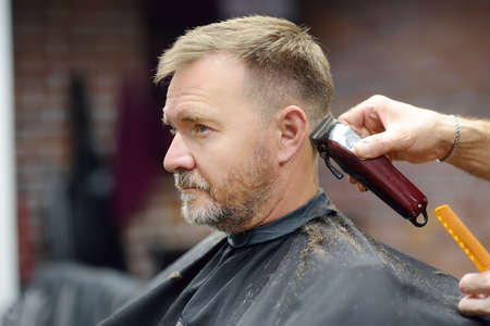 Barber master is shaving of handsome mature bearded man in salon. Hair artist making hairstyle for...