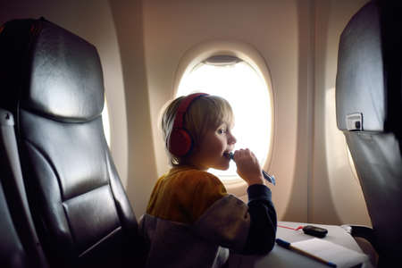 Cute little boy traveling by an airplane. Child using player to listen to a music or audiobook during the flight and eating cereal bar. Entertainment for kids on a board of planeの写真素材