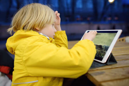 Little boy is playing computer game or watching cartoon by tablet pc in cafe. Parents are occuping...