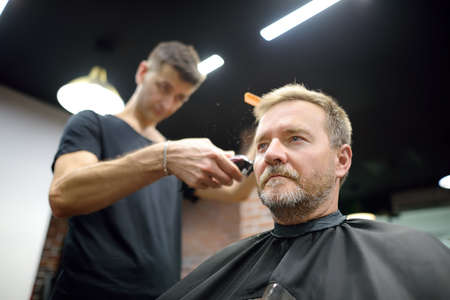 Hairdresser is cutting hair of handsome bearded mature man in salon. Stylist making hairstyle with electric shaver for person in barbershop. Services of a professional stylist. fashion haircut for menの写真素材