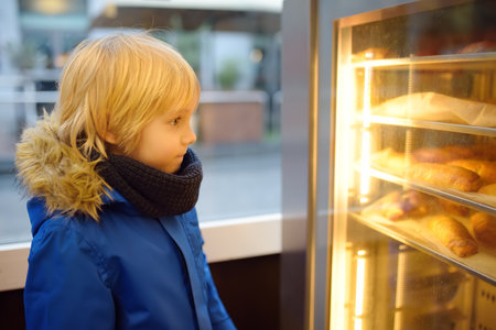 A pretty child looks at a display case with freshly baked croissants and puff pastries in a European bakery on a cold and dark winter morning. Fresh hot baked pastries in small bakeries.の写真素材