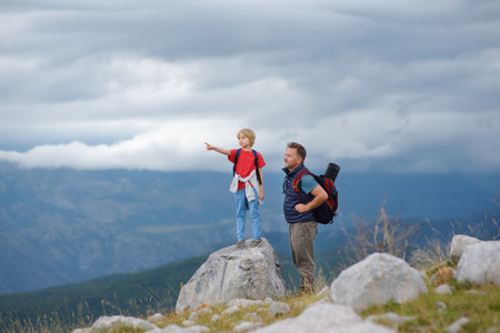 Cute schoolchild and his mature father hiking together on mountain and exploring nature. Child watching a cloud. Concepts of adventure, scouting and hiking tourism for family with kids.の写真素材