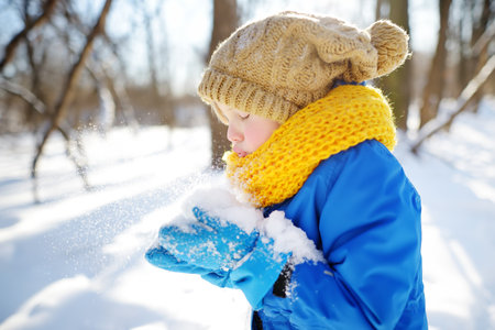 Little boy blowing snow from his hands. Child enjoy walking in the park on snowy day. Baby having fun during snowfall. Outdoor winter activities for family with kids.の写真素材