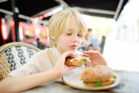 Cute blonde boy eating large bagel with salmon and arugula in outdoor fast food restaurant. Healthy meals for kids.の写真素材