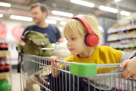 Cute preschooler boy with headphone and player is sitting in a cart at a food store or supermarket. A child is listening to music or an audiobook while his parents buying groceries. shopping with kidsの写真素材