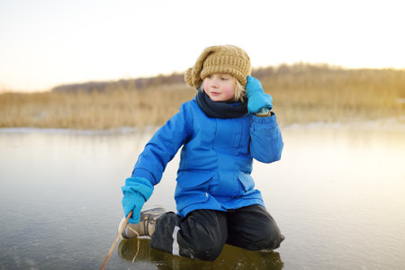 Cute preschool boy is playing on the ice of a frozen lake or river on a cold sunny winter sunset. Child having fun with icicle and dry reed plant during family hiking. Kids outdoor games in winterの写真素材