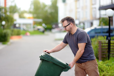 Mature man pulling out a large green plastic garbage container in front of the townhouse to the roadway of the street. Garbage collection and recycling trash, the zero waste concept. Waste managementの写真素材