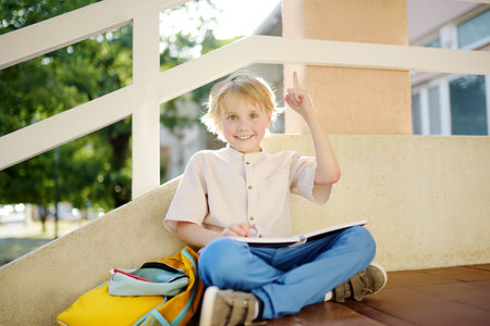 Little student doing homework on break on stair of elementary school building. Cute boy having idea...