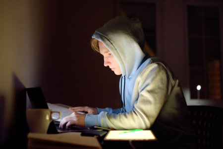 Concentrated teenager studying late night at home office with laptop and tablet. Modern technologies...