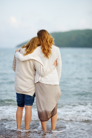 Redhead senior mother and her adult daughter are walking together on the sea shore. Happy loving mom and her grown up child embracing and admiring of nature.の写真素材