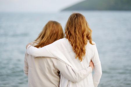 Redhead senior mother and her adult daughter are walking together on the sea shore. Happy loving mom and her grown up child embracing and admiring of nature.の写真素材