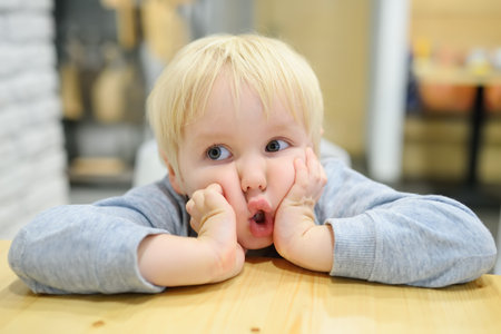 Funny emotional child is at a table in a cafe. Cute little boy sitting in restaurant and waiting for...