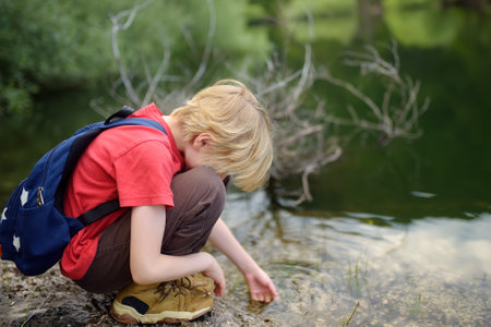 Preteen boy in red shirt is exploring nature and playing with water in lake during hiking in mountains valley. Active leisure for energetic children. Summer camp outdoor recreation for kidsの写真素材