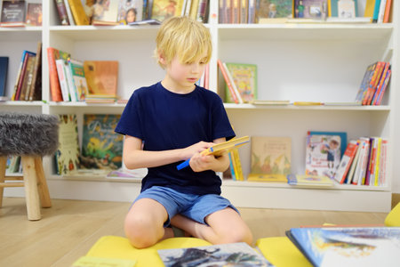 A preteen boy leafing through a book while sitting at the bookshelves in a school library or bookstore. Smart kid reading comics or adventure bookの写真素材