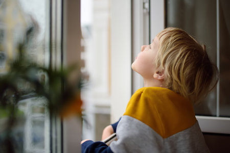 Little boy watching the rain outside at opened window. A child of primary school age is at risk of falling from a window from a great height. Dangers in the home for a small kidの写真素材