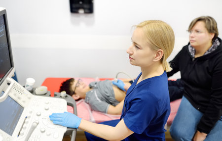 Doctor making abdominal ultrasound for boy using scanner machine. Female doc runs ultrasound sensor over patient tummy looking at screen. Mother supports son during examination of internal organsの写真素材