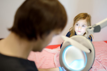 A caring doctor checks moles on the skin of a small child using lamp with magnifying glass. A dermatologist looks at a rash on the leg of a girl. Baby with mom at a pediatrician appointmentの写真素材