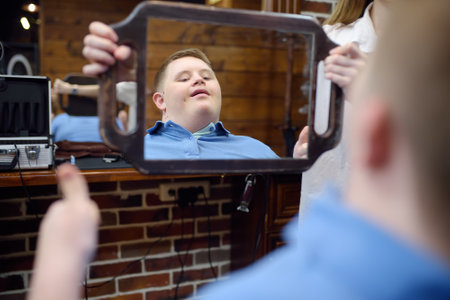 Portrait of a teenager with Down syndrome in a barbershop. Boy with Down syndrome smiles looking at himself in the mirror after a haircutの写真素材